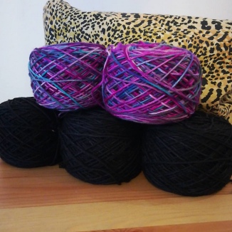 Two skeins of purple and green yarn resting atop three skeins of black yarn in front of a leopard print fabric bag.